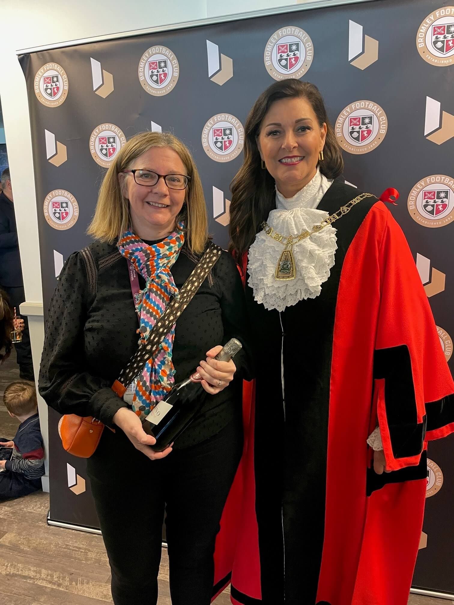 Two women smiling, one in a black outfit holding a bottle, the other in ceremonial robes, standing in front of a banner. - Home Instead
