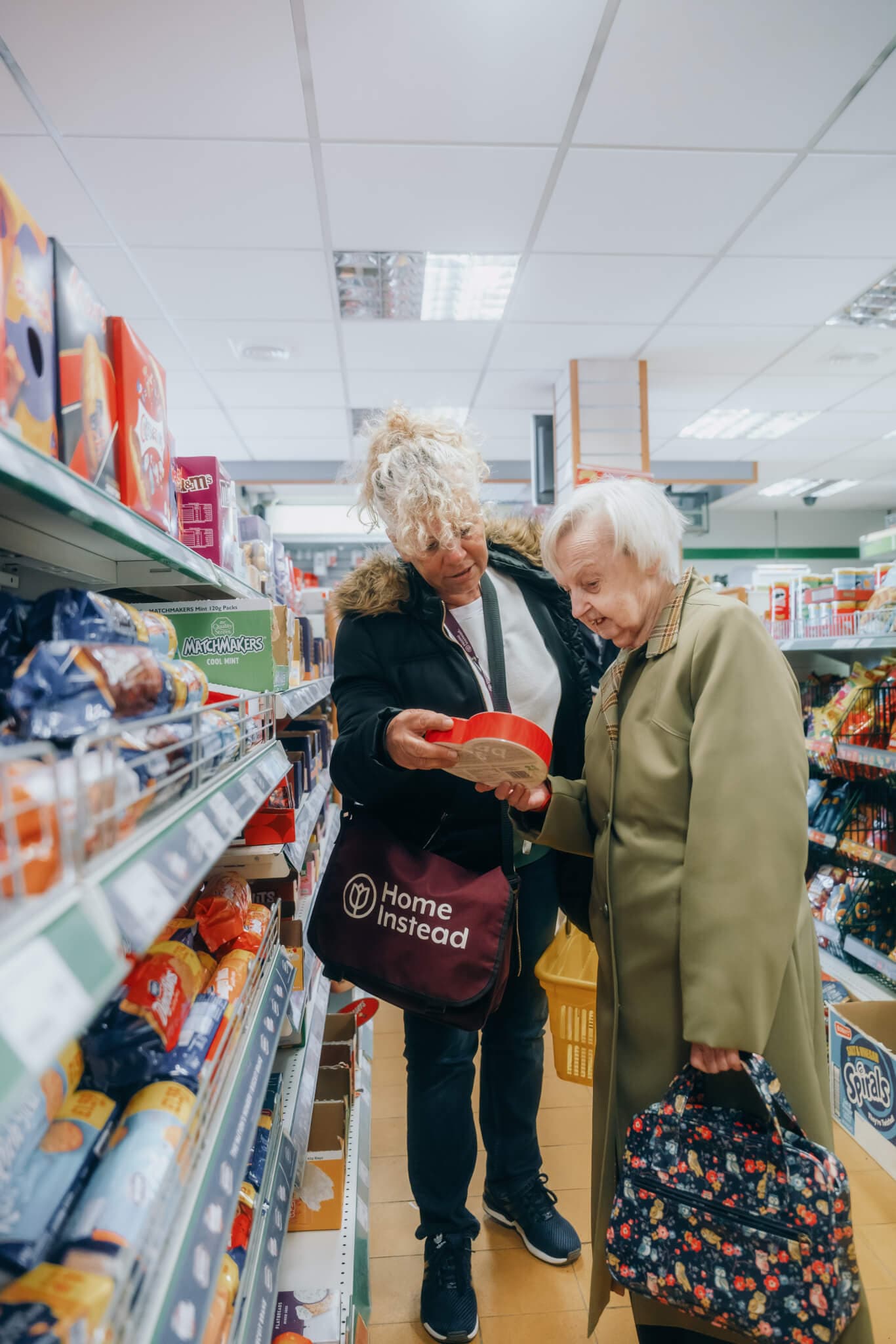 A caregiver assists an elderly woman in choosing an item from a grocery store shelf. - Home Instead