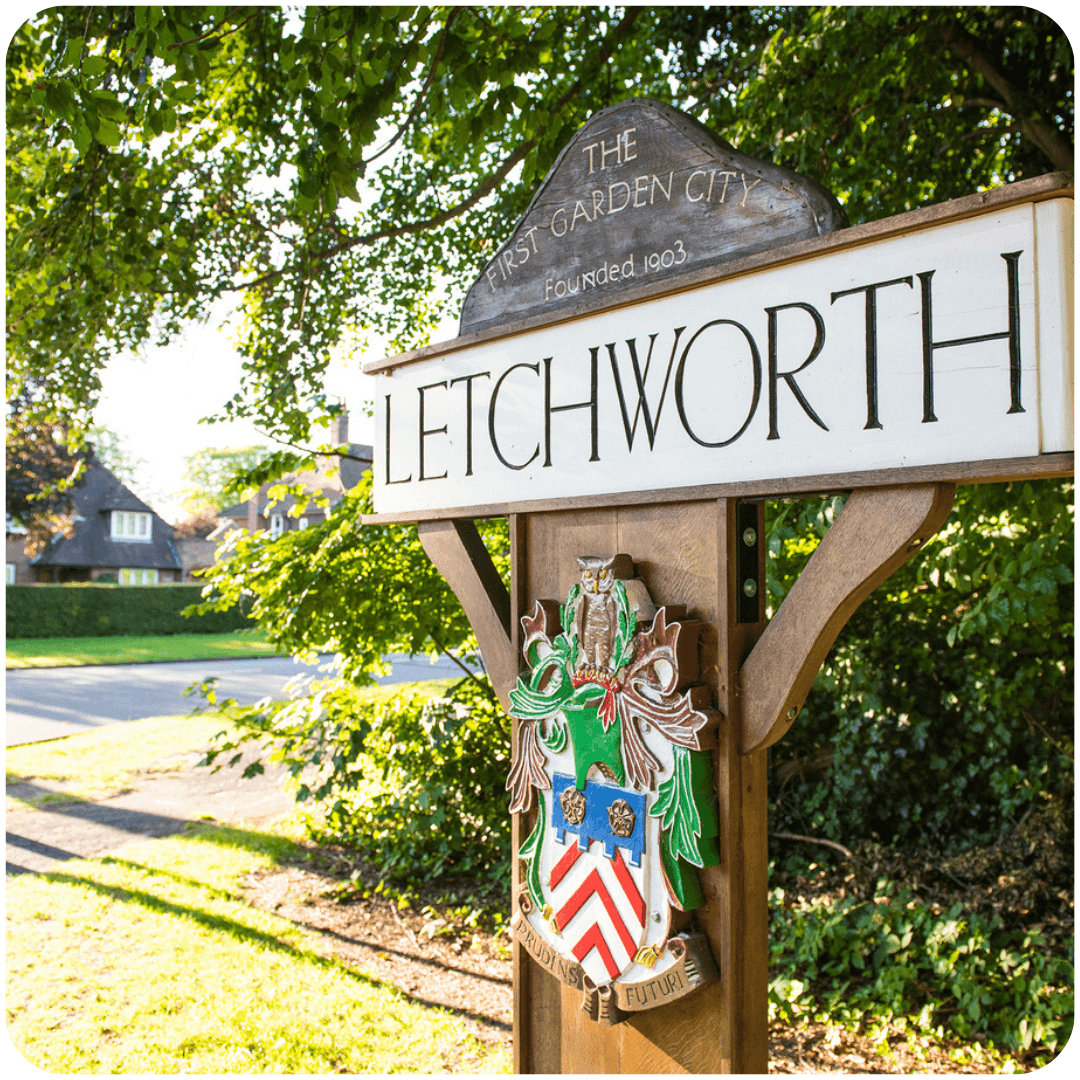 Sign for the town of Letchworth, "The First Garden City," with a coat of arms, surrounded by trees and houses. - Home Instead