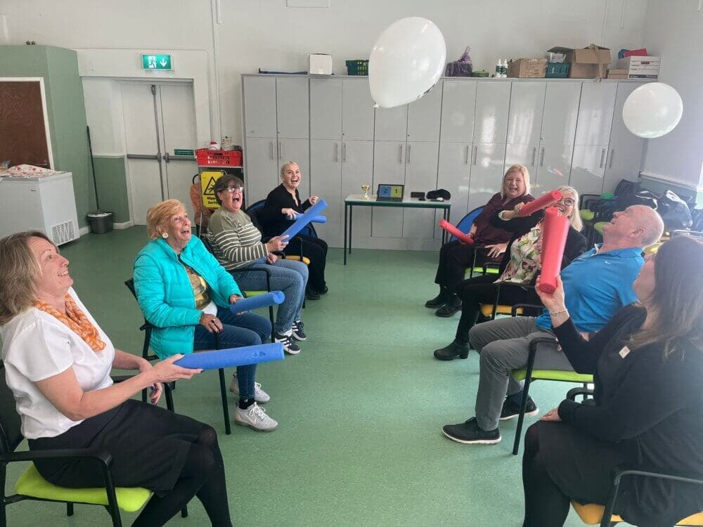 A group of people sitting in a circle, smiling and playing with red and blue pool noodles, with a balloon in the air. - Home Instead