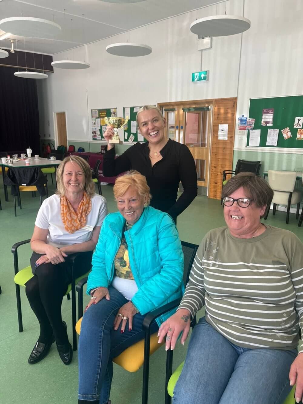 Four women smiling and sitting in a bright room, one woman standing behind the others holding up a card. - Home Instead