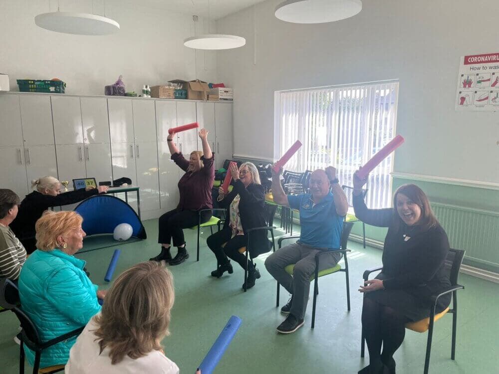 A group of people sitting in chairs, holding foam sticks, and participating in a lively indoor activity session. - Home Instead