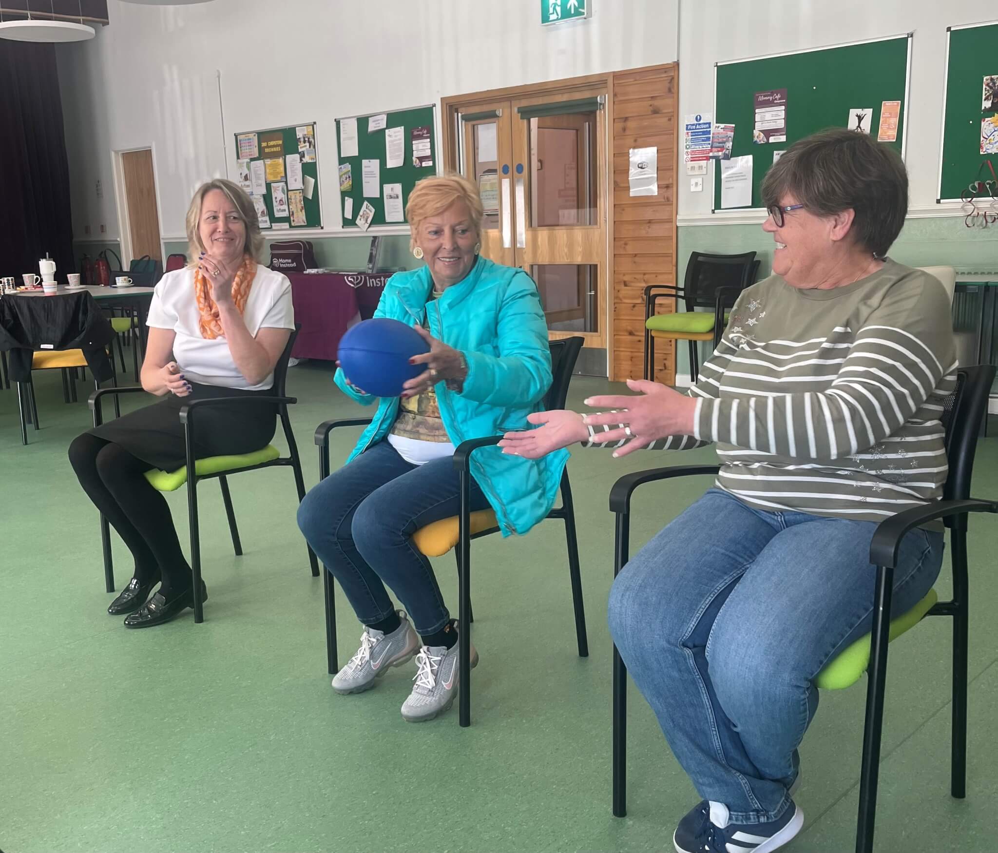 Three women sit in chairs in a community center, passing a blue ball and smiling. Bulletin boards are behind them. - Home Instead