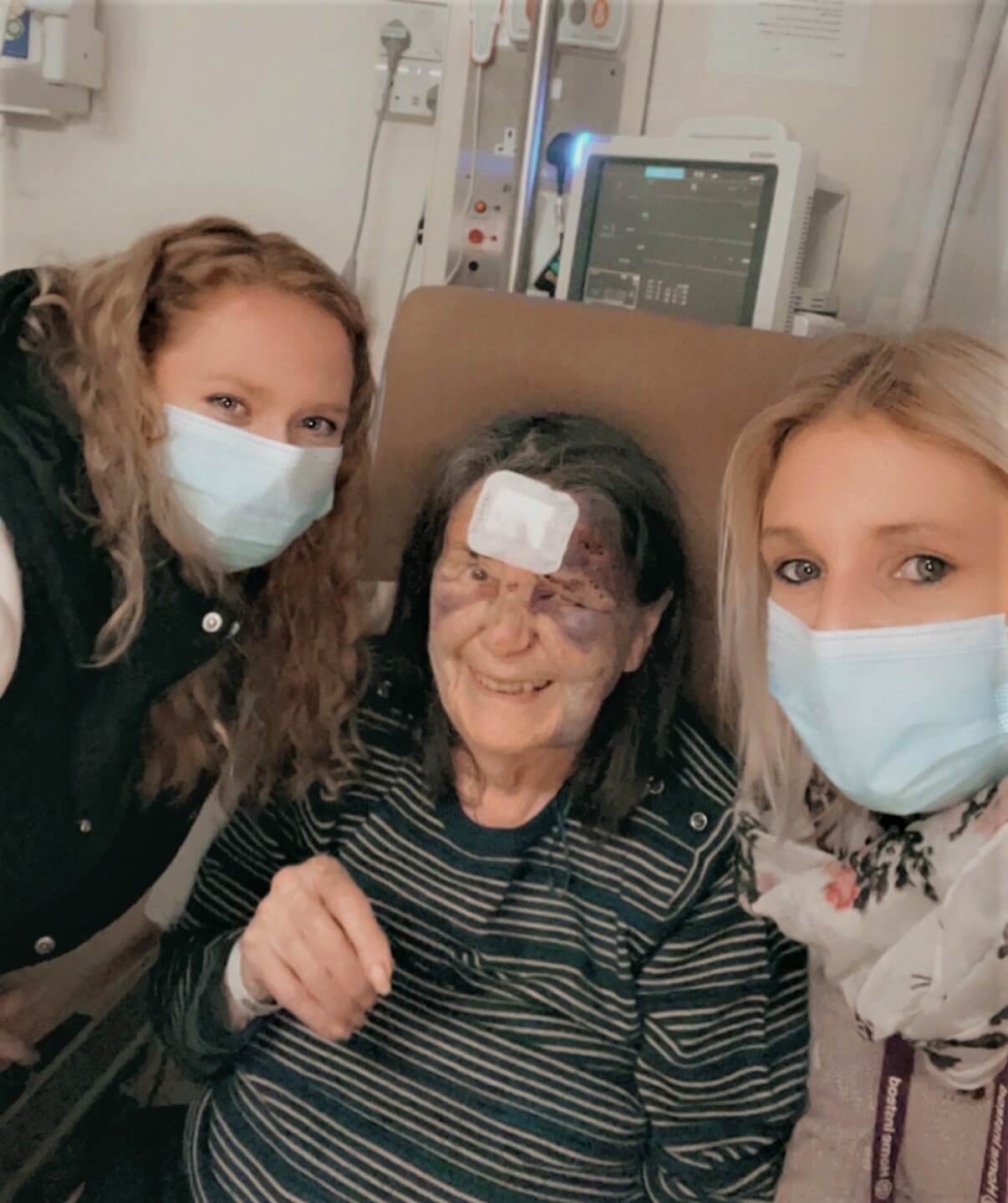 Three women, two standing and one seated with a head injury, wearing masks, posing for a photo in a medical setting. - Home Instead
