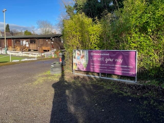 A banner for "Home Instead" on a fence beside a dirt path, with buildings and trees in the background. - Home Instead