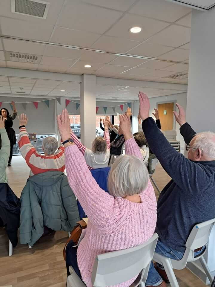 A group of elderly people seated in a room performing chair exercises with their hands raised. - Home Instead