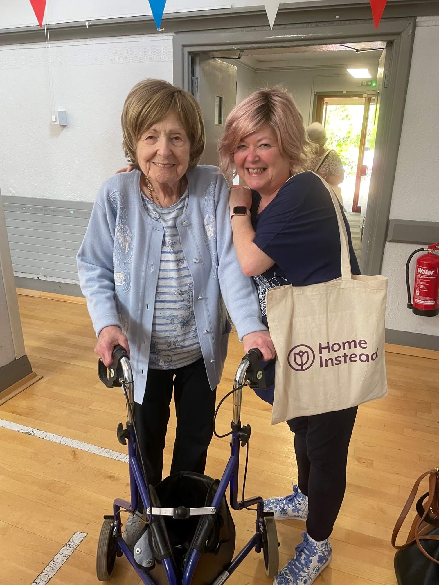 Two women smile together, one elderly using a walker and the other with an arm around her, holding a "Home Instead" bag. - Home Instead