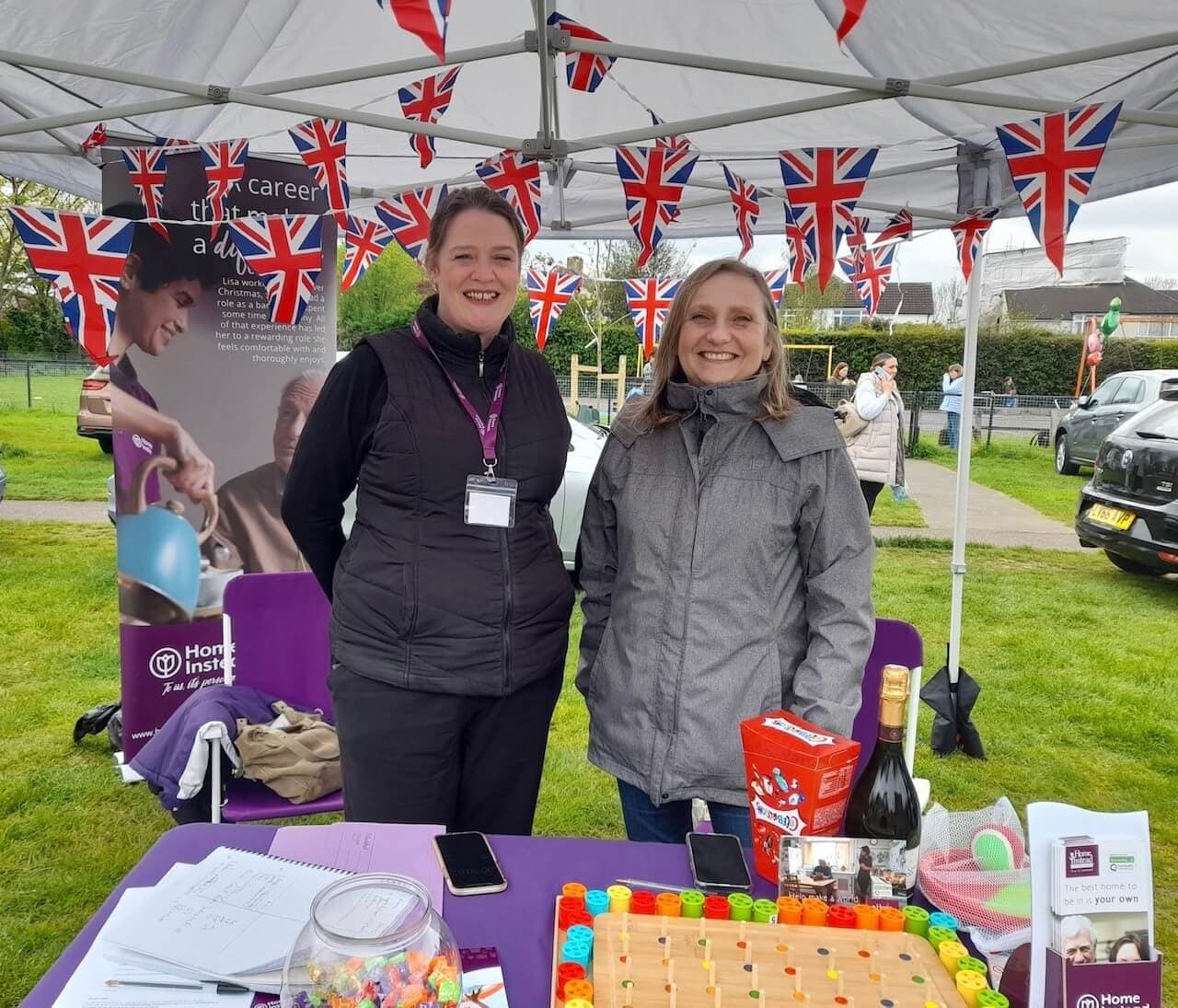 Two women smiling at a festival booth decorated with Union Jack flags, standing behind a table with various items on it. - Home Instead