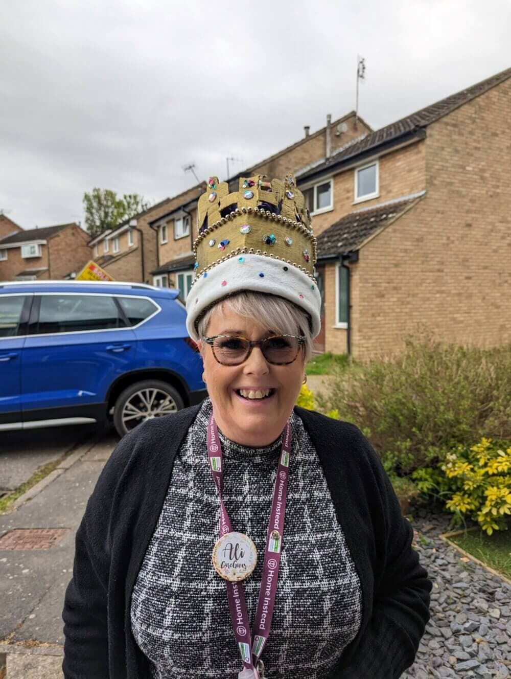 Person smiling and wearing a crown-shaped hat, with a lanyard and badge, standing in a residential area with houses and a blue car. - Home Instead