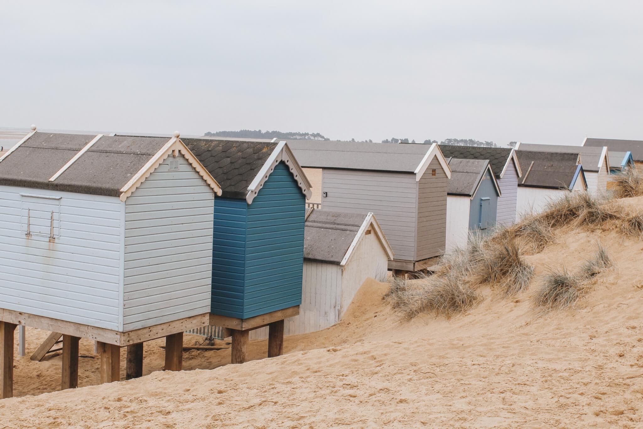 A row of colorful beach huts on sandy dunes, with a cloudy sky in the background. - Home Instead