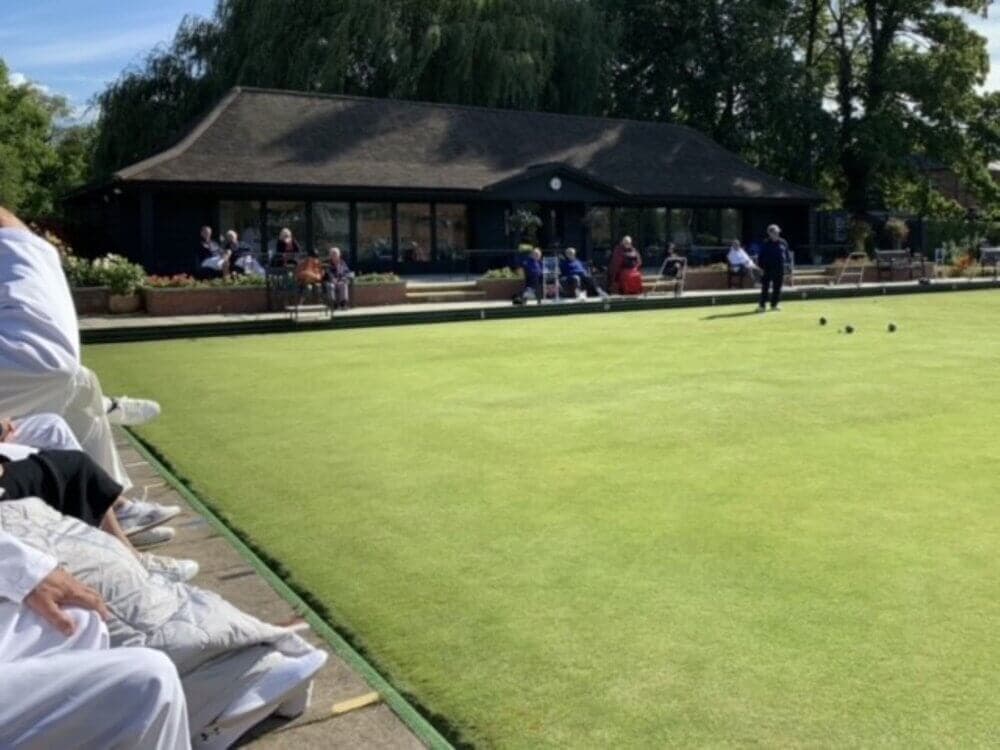 People playing lawn bowling on a sunny day with a clubhouse and spectators in the background. - Home Instead