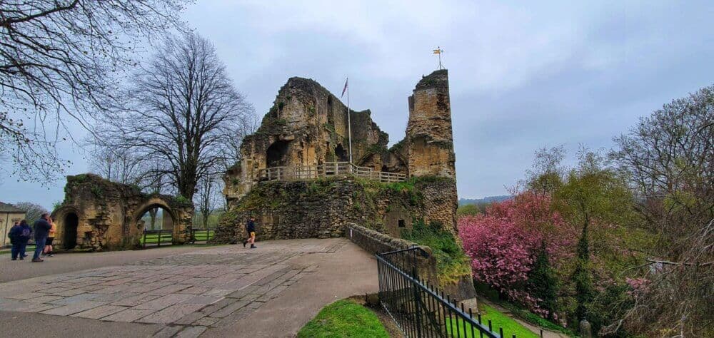 Ruins of an ancient stone castle with flags, surrounded by trees and a pathway, under a cloudy sky. - Home Instead