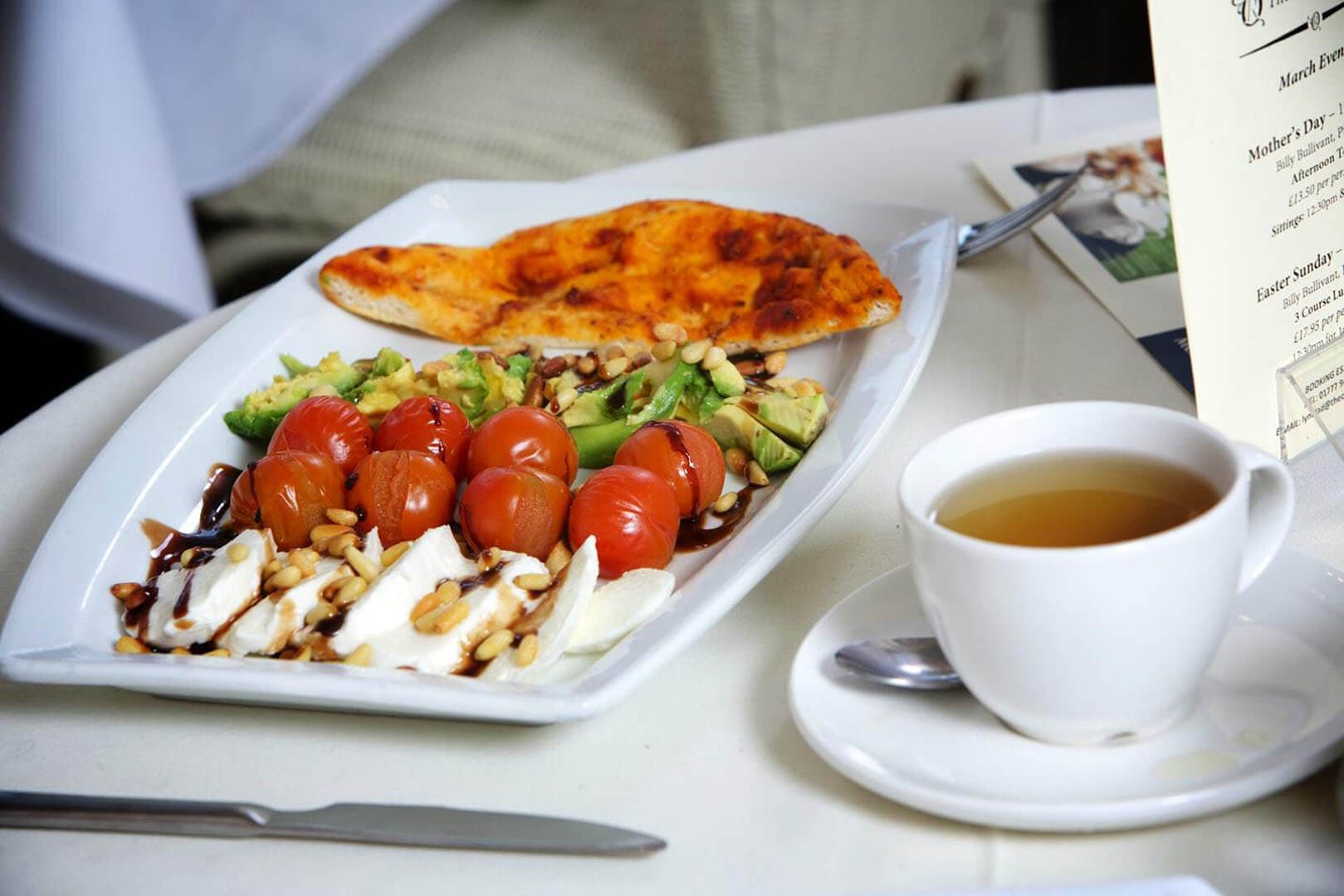 A plate of sliced tomatoes, avocado, mozzarella, and flatbread with a cup of tea on a white table. - Home Instead