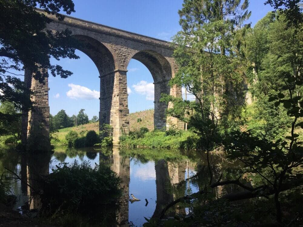 Stone arch bridge over a calm river surrounded by lush greenery and trees, with a clear blue sky in the background. - Home Instead