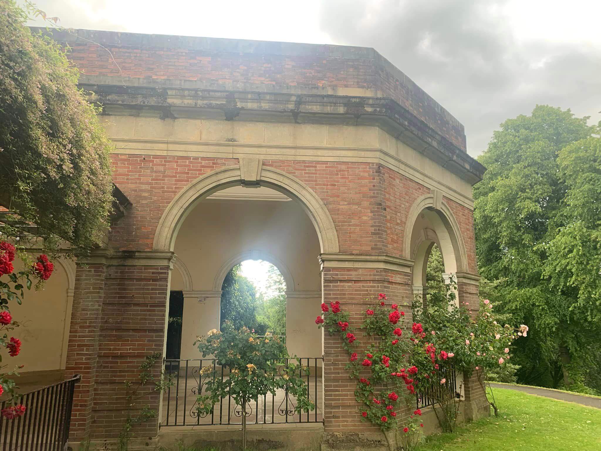 A brick pavilion with archways is adorned with red roses, set against a cloudy sky and green surroundings. - Home Instead
