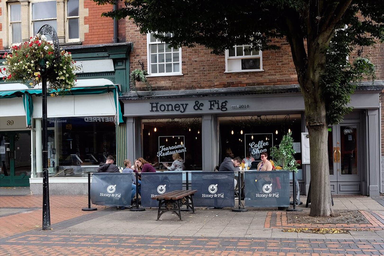 People dining outdoors at Honey & Fig restaurant and coffee shop on a quaint street. Flowers hanging from lamppost. - Home Instead