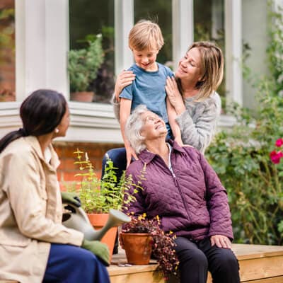 A woman embraces a child while speaking with an older woman sitting on a step, another woman nearby holds a watering can. - Home Instead