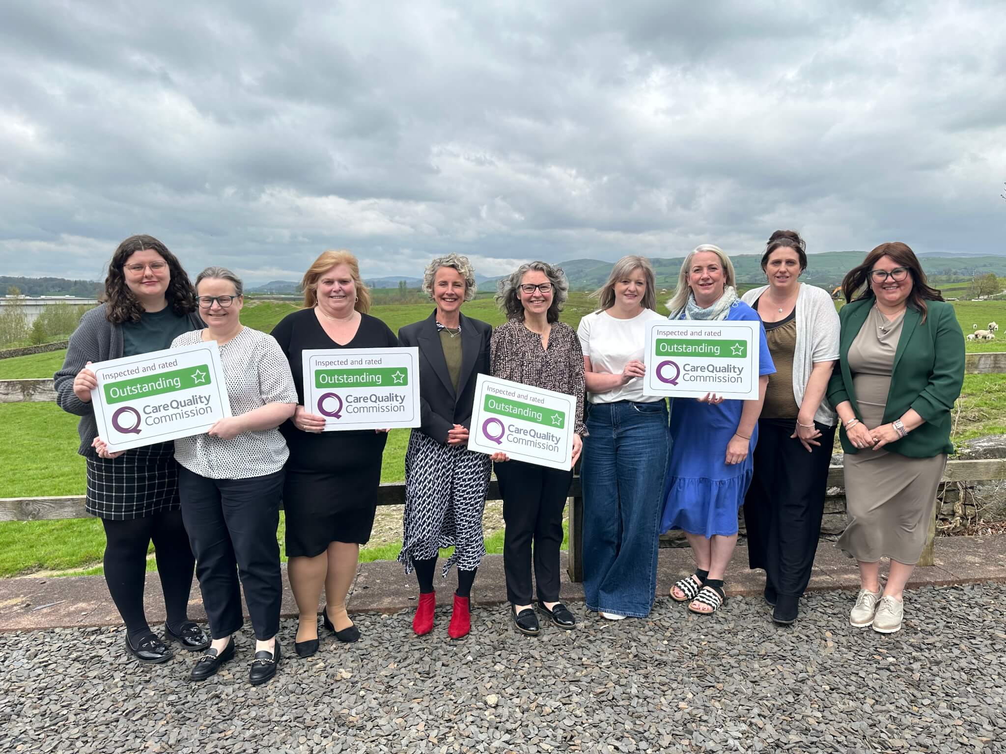 A group of people standing outdoors, holding "Outstanding" ratings from the Care Quality Commission. - Home Instead