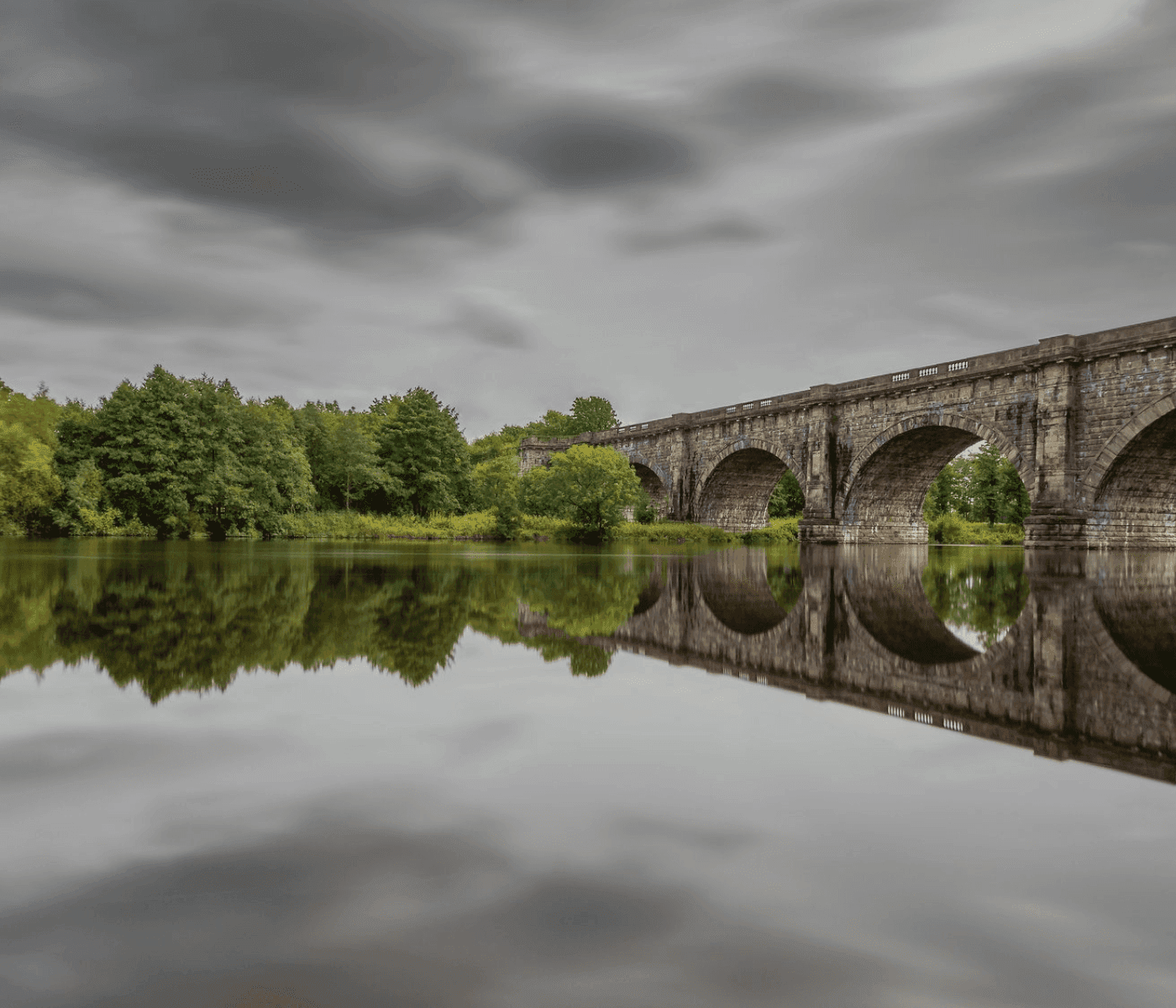 Stone bridge with arches reflected in a calm river, surrounded by lush greenery under a cloudy sky. - Home Instead