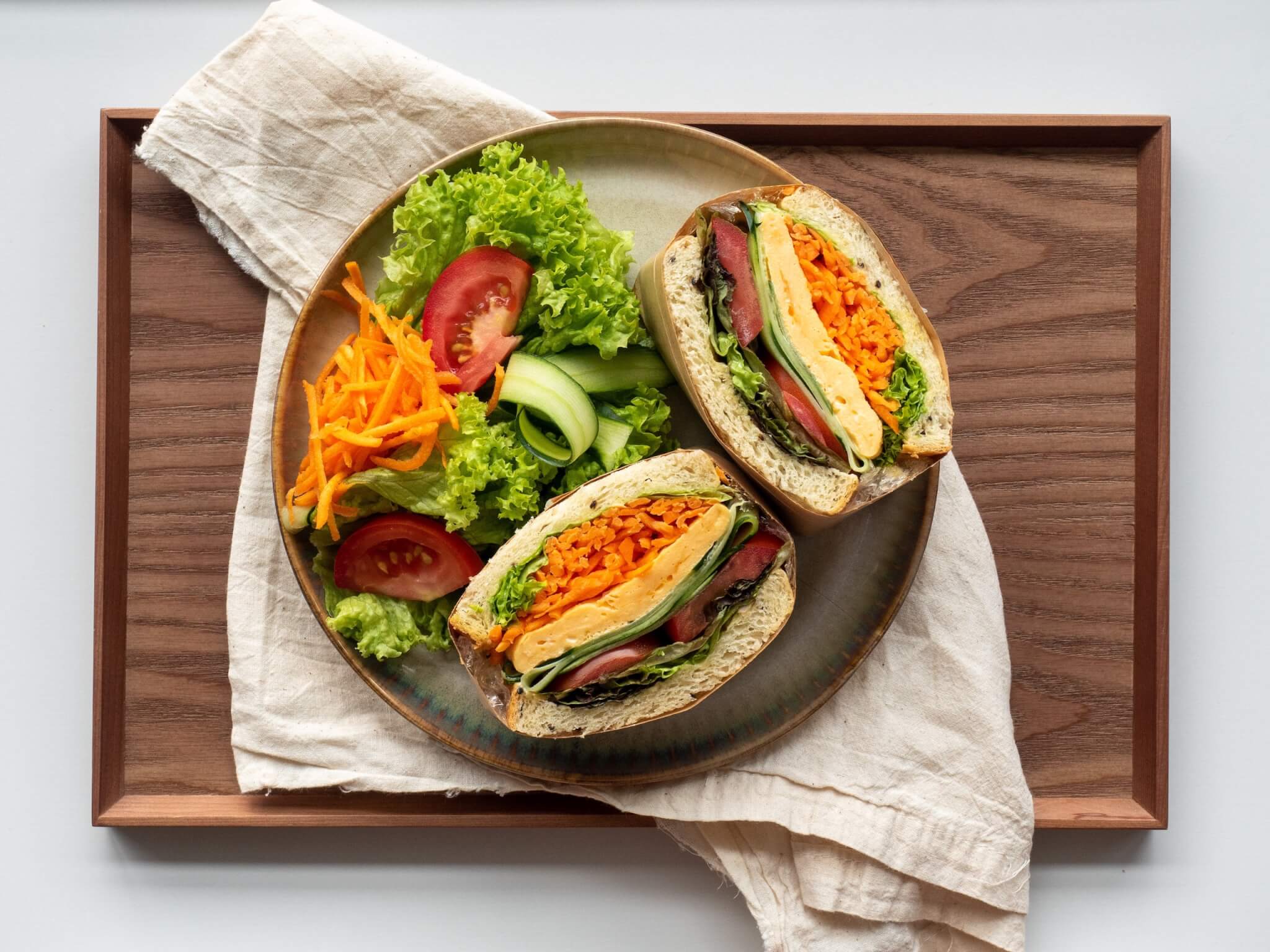 Vegetable sandwich with lettuce, tomato, and carrots on a wooden tray beside fresh vegetable salad on a plate. - Home Instead