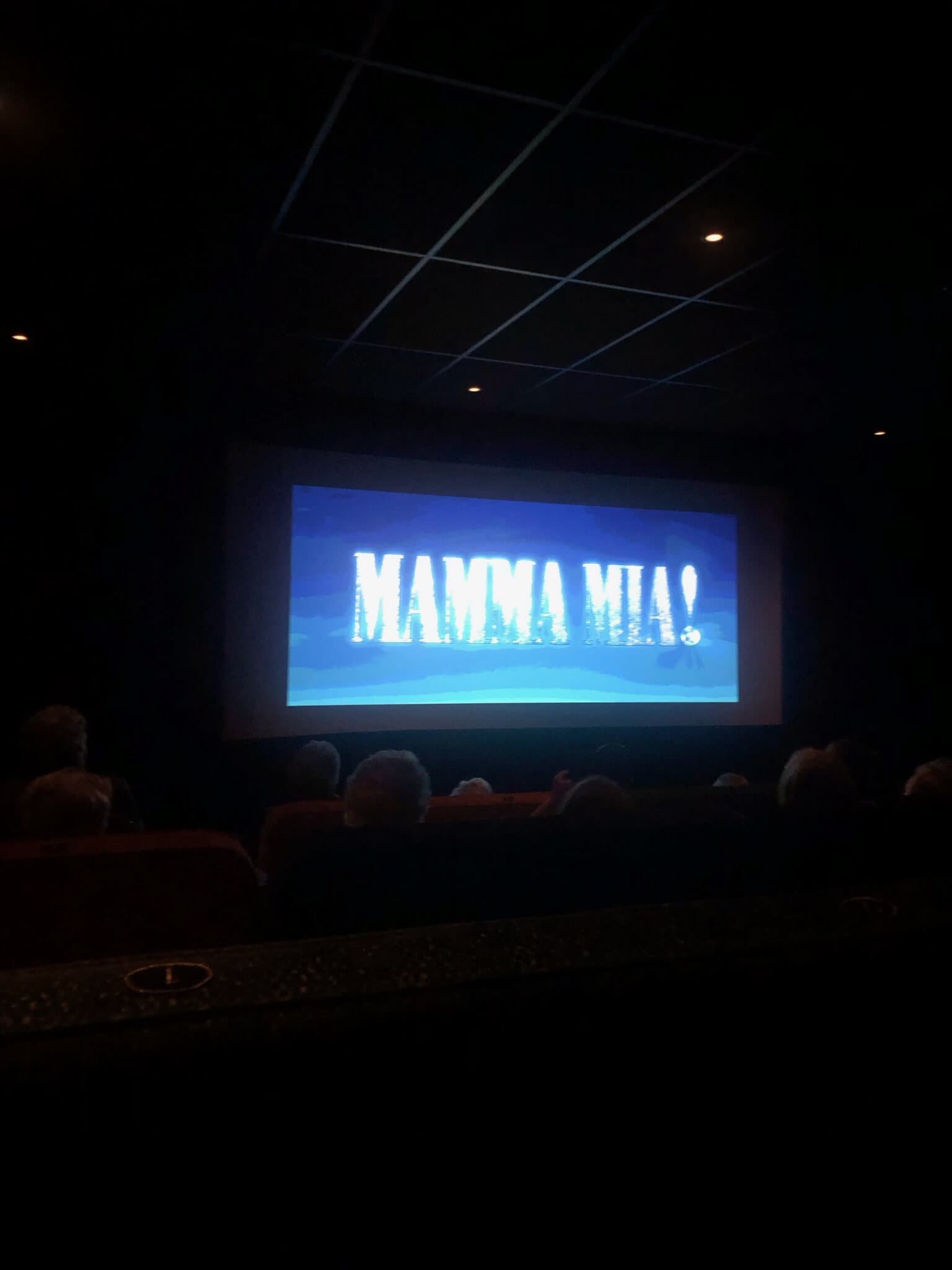 Movie theater with an audience watching the title screen of "Mamma Mia!" on the big screen. - Home Instead
