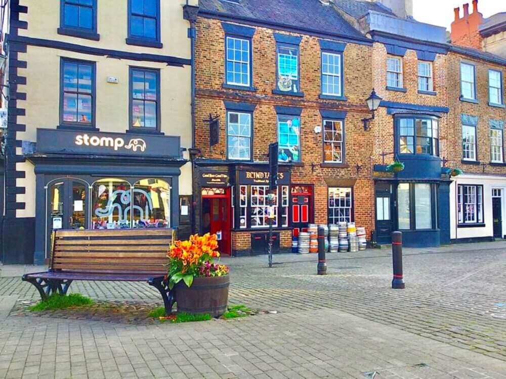 A street with historic brick buildings, a bench, a flower arrangement, and a pub named "Stomp". - Home Instead