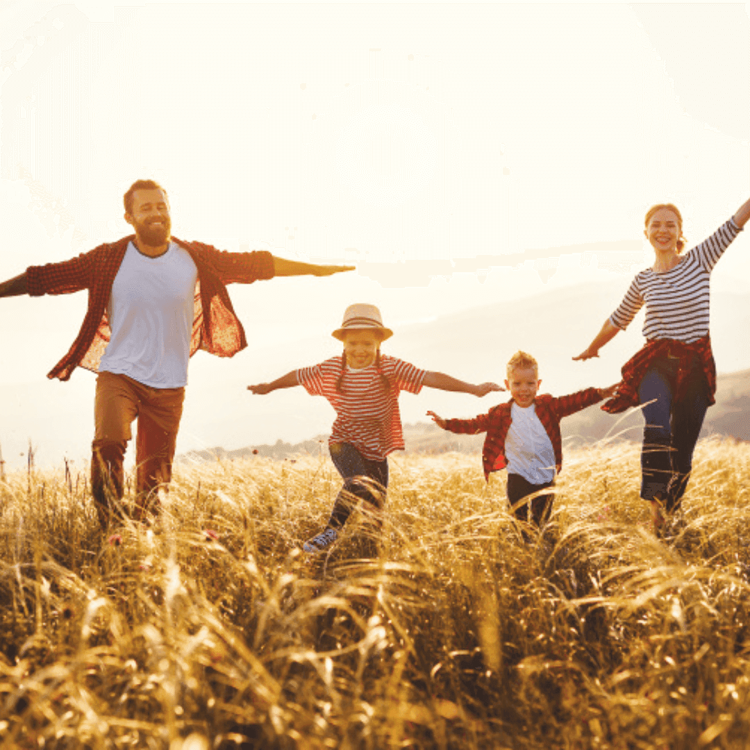 A family of four, two adults and two children, running through a field with their arms outstretched, smiling joyfully in the sunlight. - Home Instead