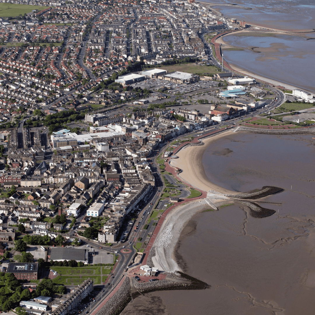 Aerial view of a coastal town with a sandy beach, numerous buildings, roads, and a receding tide exposing mudflats. - Home Instead