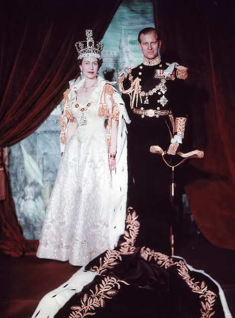 A crowned woman in a white robe stands beside a man in black uniform adorned with medals and tassels, in front of red curtains. - Home Instead