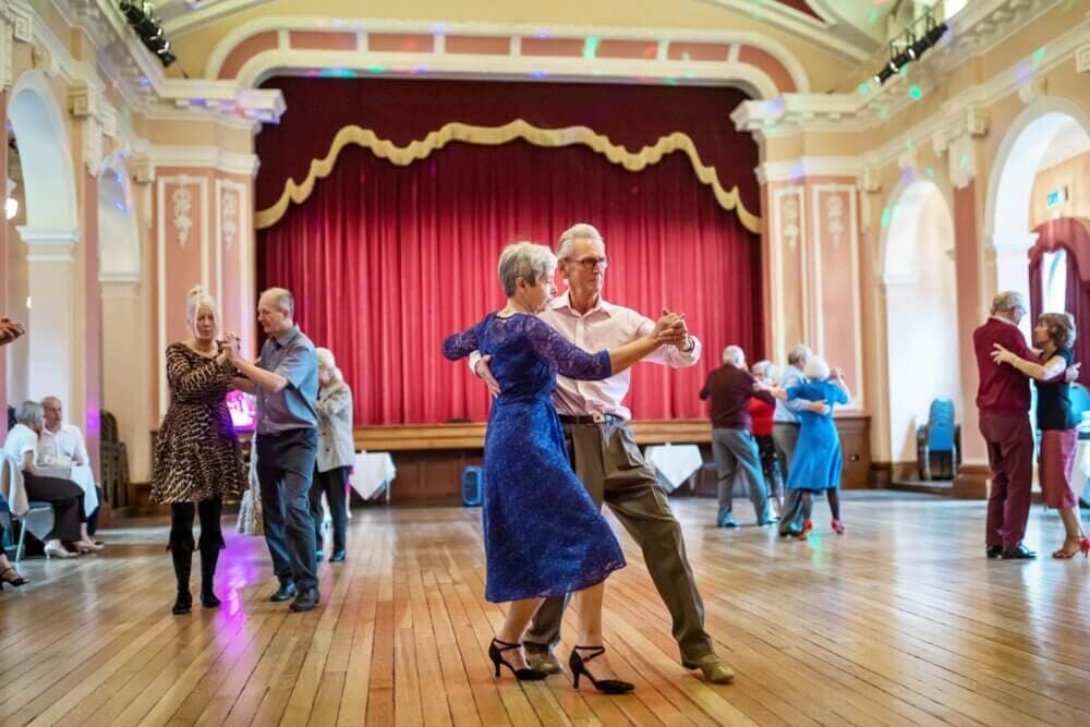 A group of people ballroom dancing in a grand hall with a red curtain and arched windows. - Home Instead