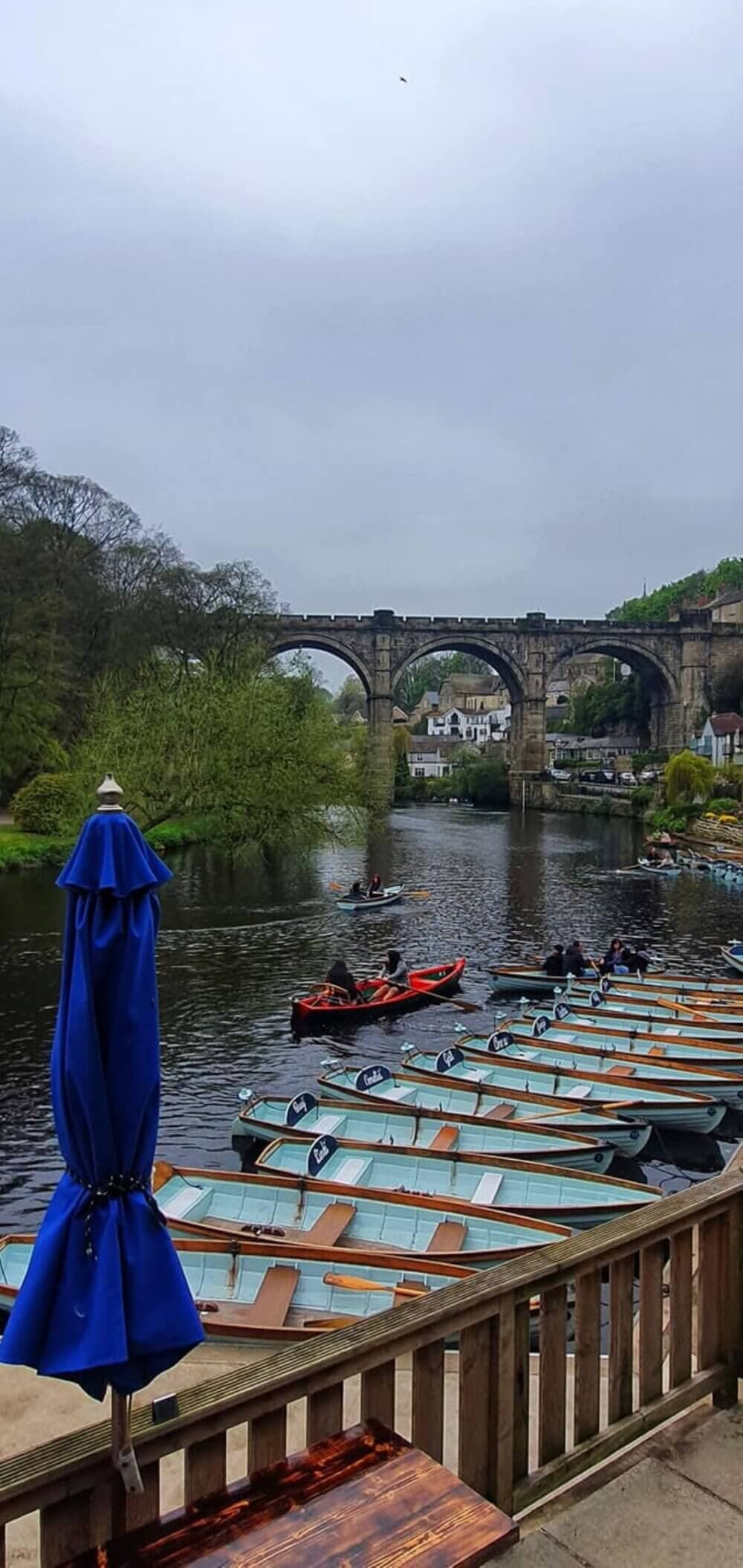 A riverside view with boats lined up on the water, an old stone bridge in the background, and a blue umbrella in the foreground. - Home Instead