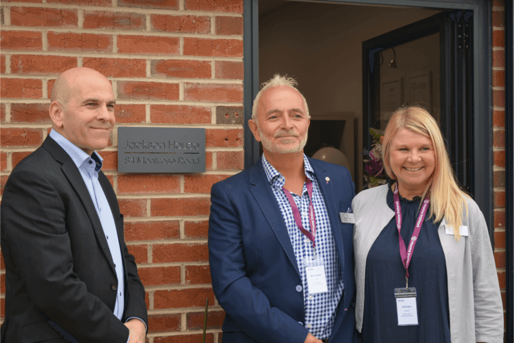 Three people standing in front of a brick building with a sign that reads "Jackson House, 3A Montrose Road. - Home Instead