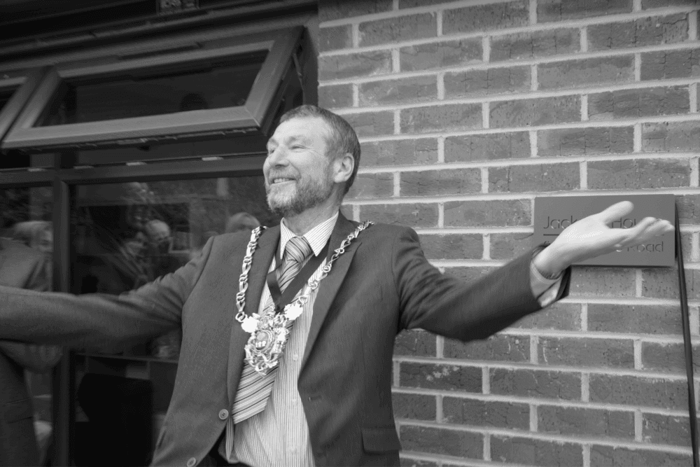 A man wearing a suit and a ceremonial chain, gestures with open arms in front of a brick wall. - Home Instead