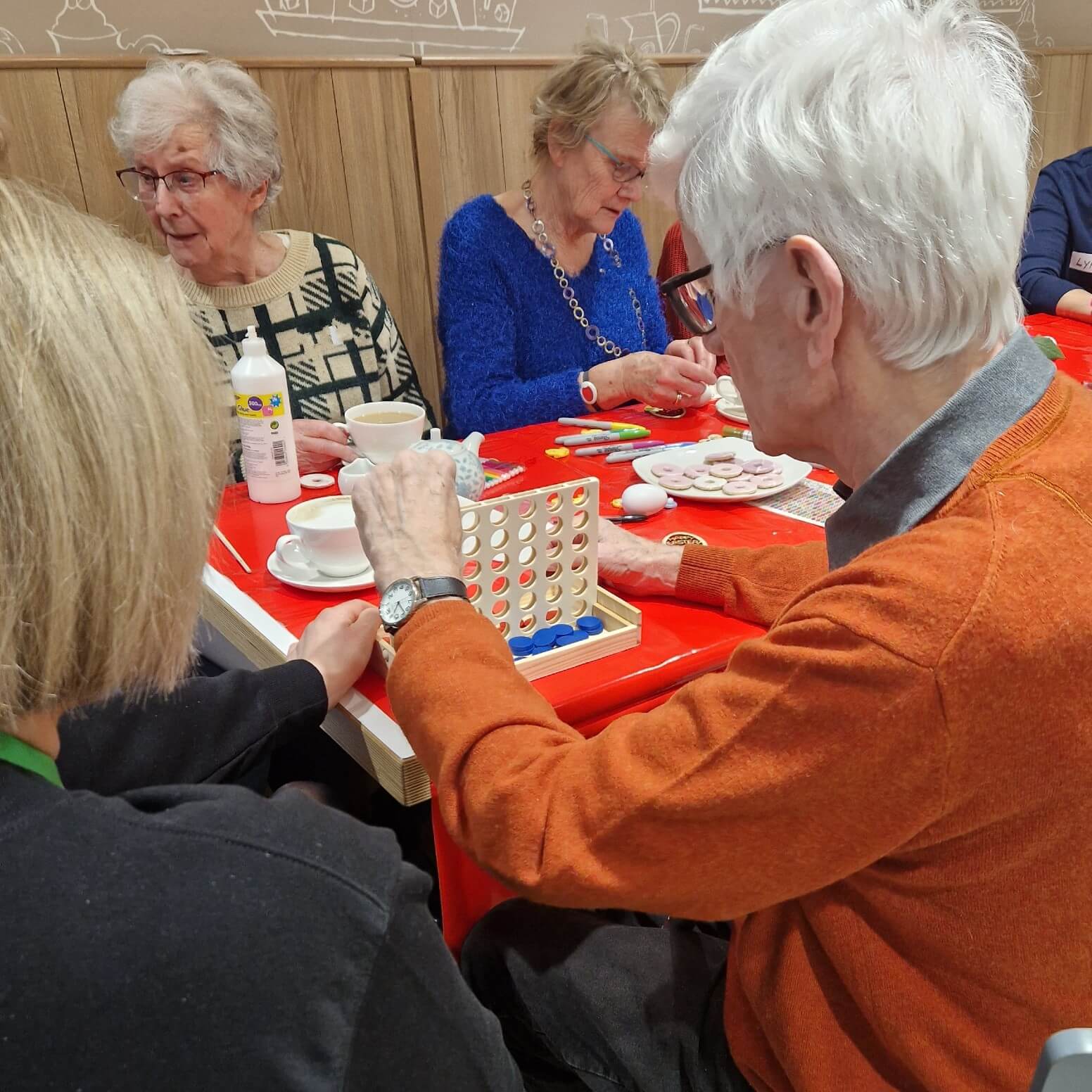 Seniors enjoying a game of Connect 4 and conversation at a social gathering with tea and snacks. - Home Instead