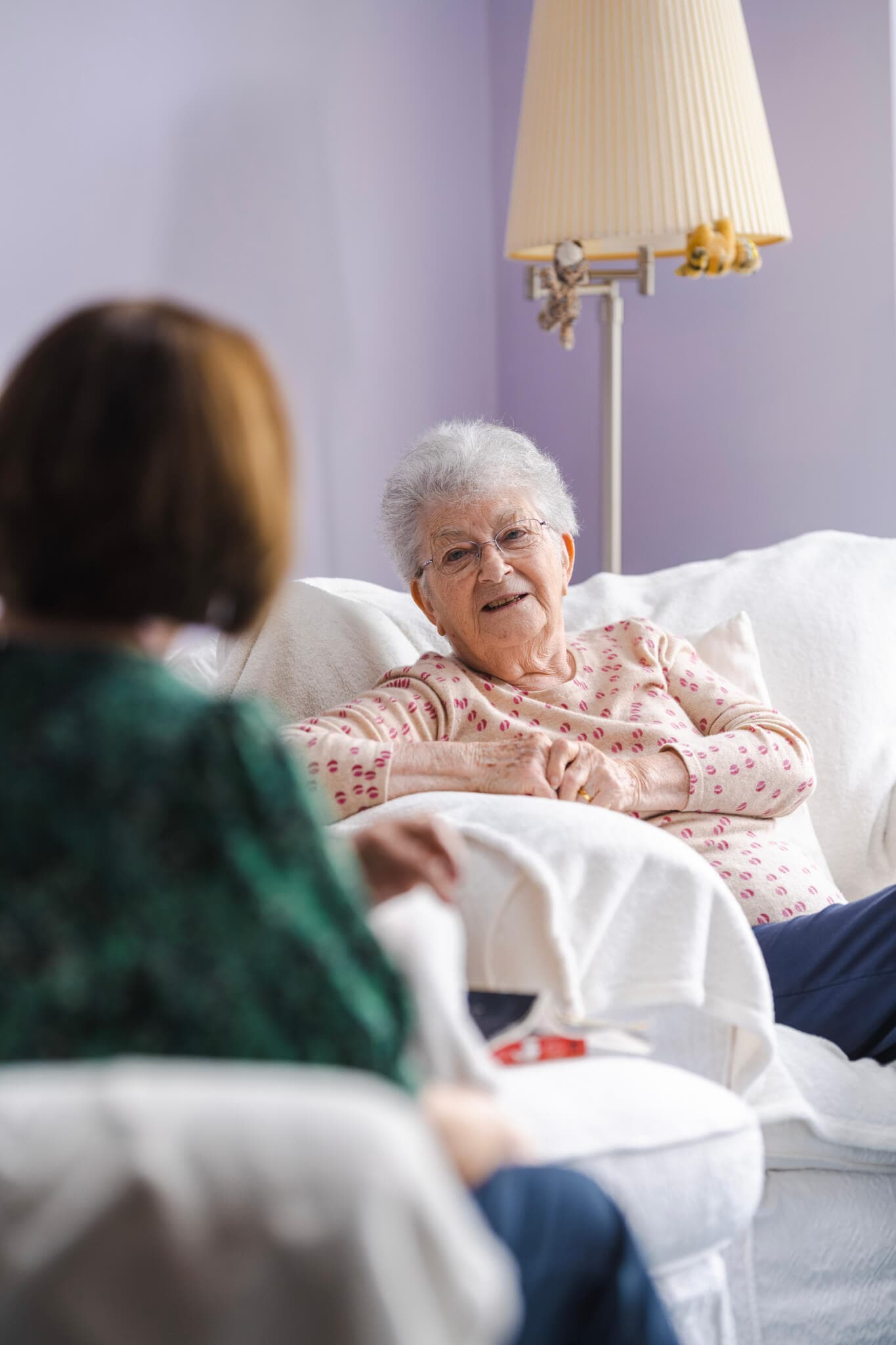 An elderly woman, smiling and sitting on a white couch, converses with another person in a green top. - Home Instead