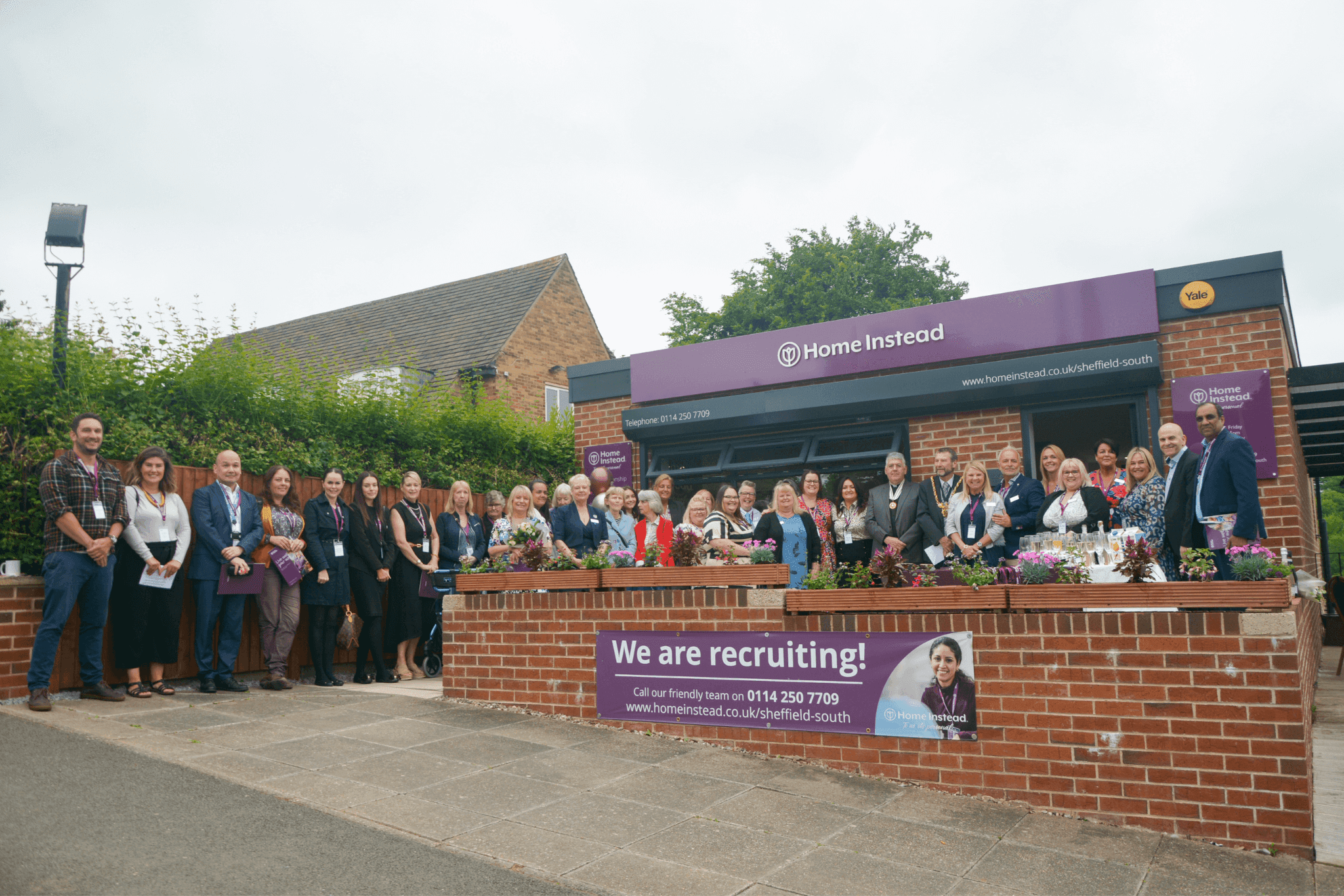 Group of people posing in front of a "Home Instead" building with a banner that reads "We are recruiting!. - Home Instead
