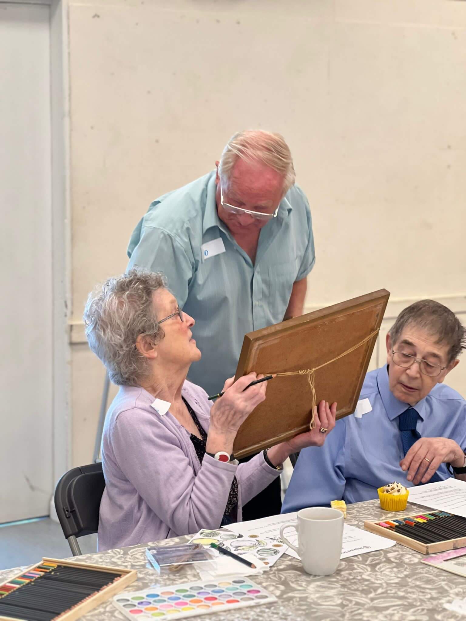 Two elderly people at a table painting, while another person stands beside them, looking at their artwork. - Home Instead