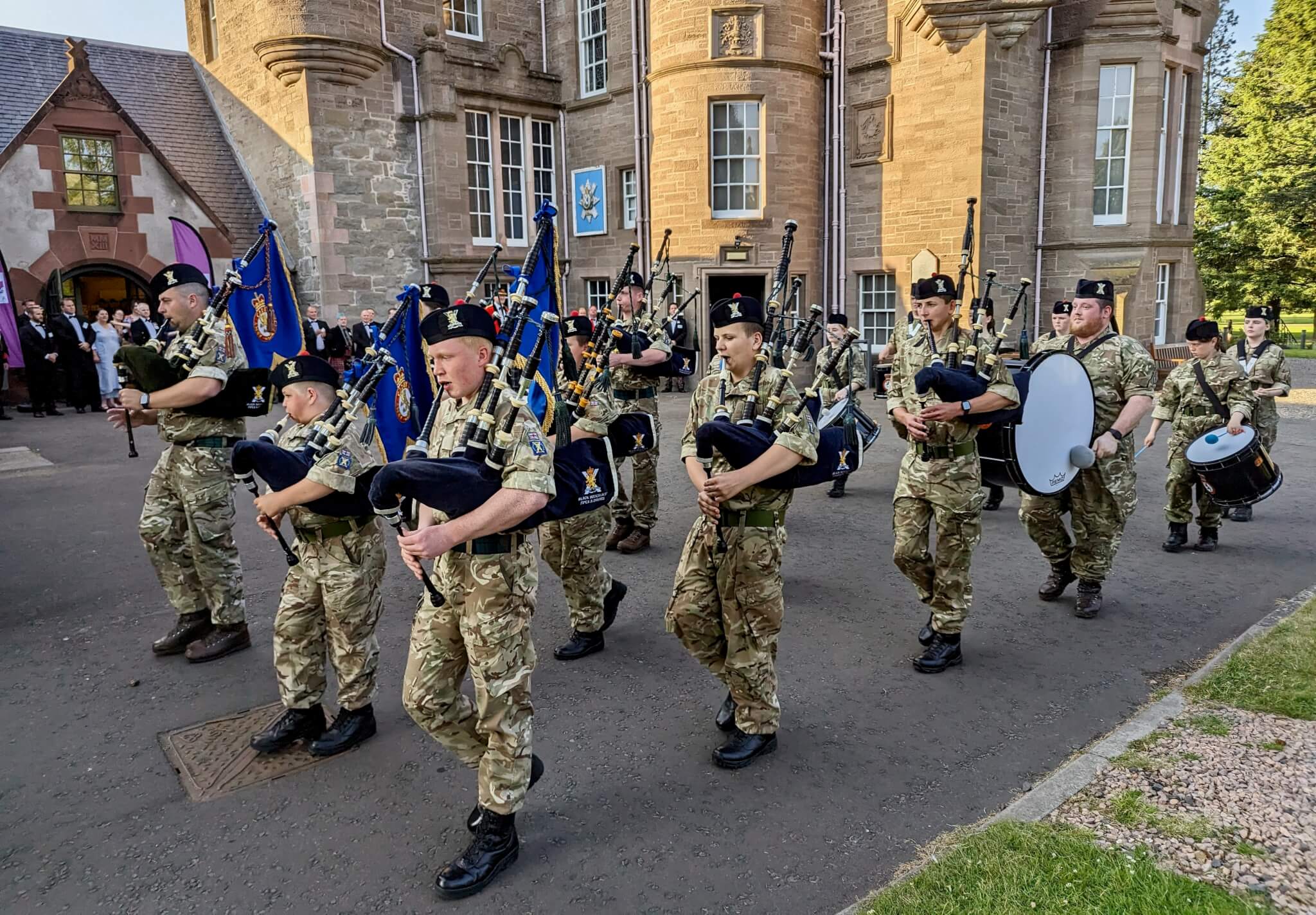 A ceremonial military band in camouflage uniforms plays bagpipes and drums while marching outside a historic stone building. - Home Instead