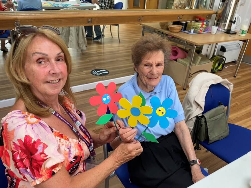Two women smile while holding colorful paper flowers in a room with wooden floors and tables. - Home Instead