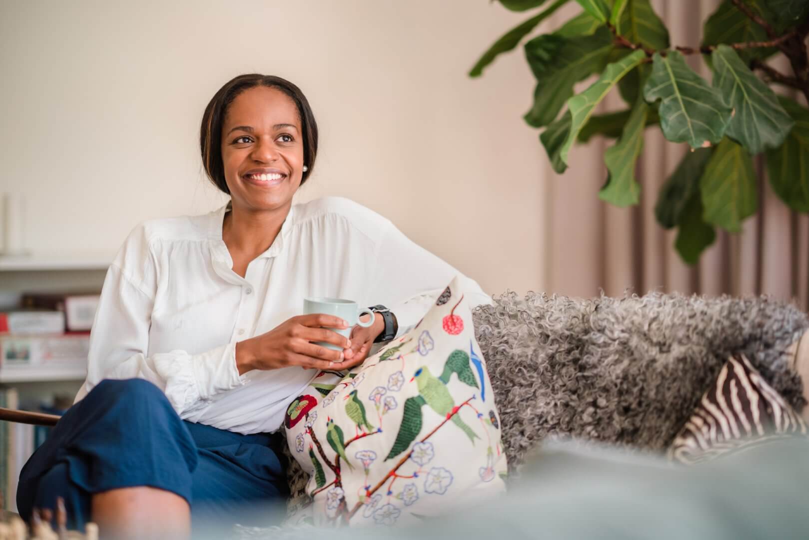 A smiling woman in a white blouse holds a mug while sitting on a couch with colorful cushions and a plant beside her. - Home Instead