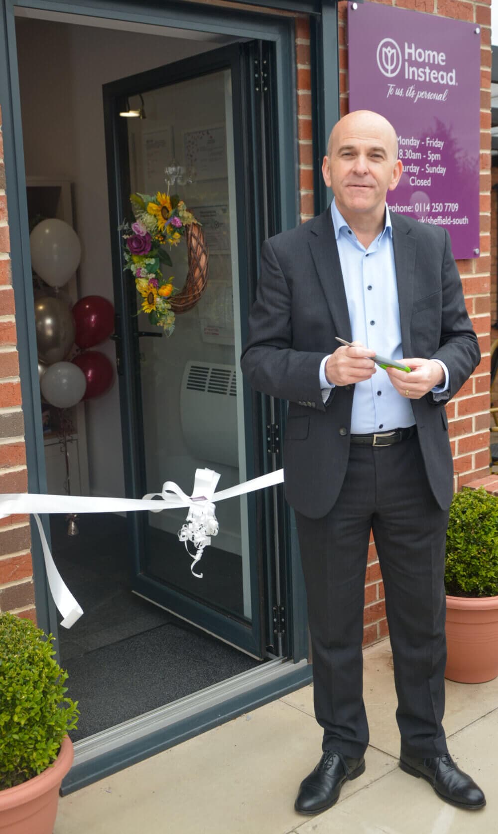 A man stands outside a door holding scissors about to cut a ribbon at an opening ceremony. - Home Instead