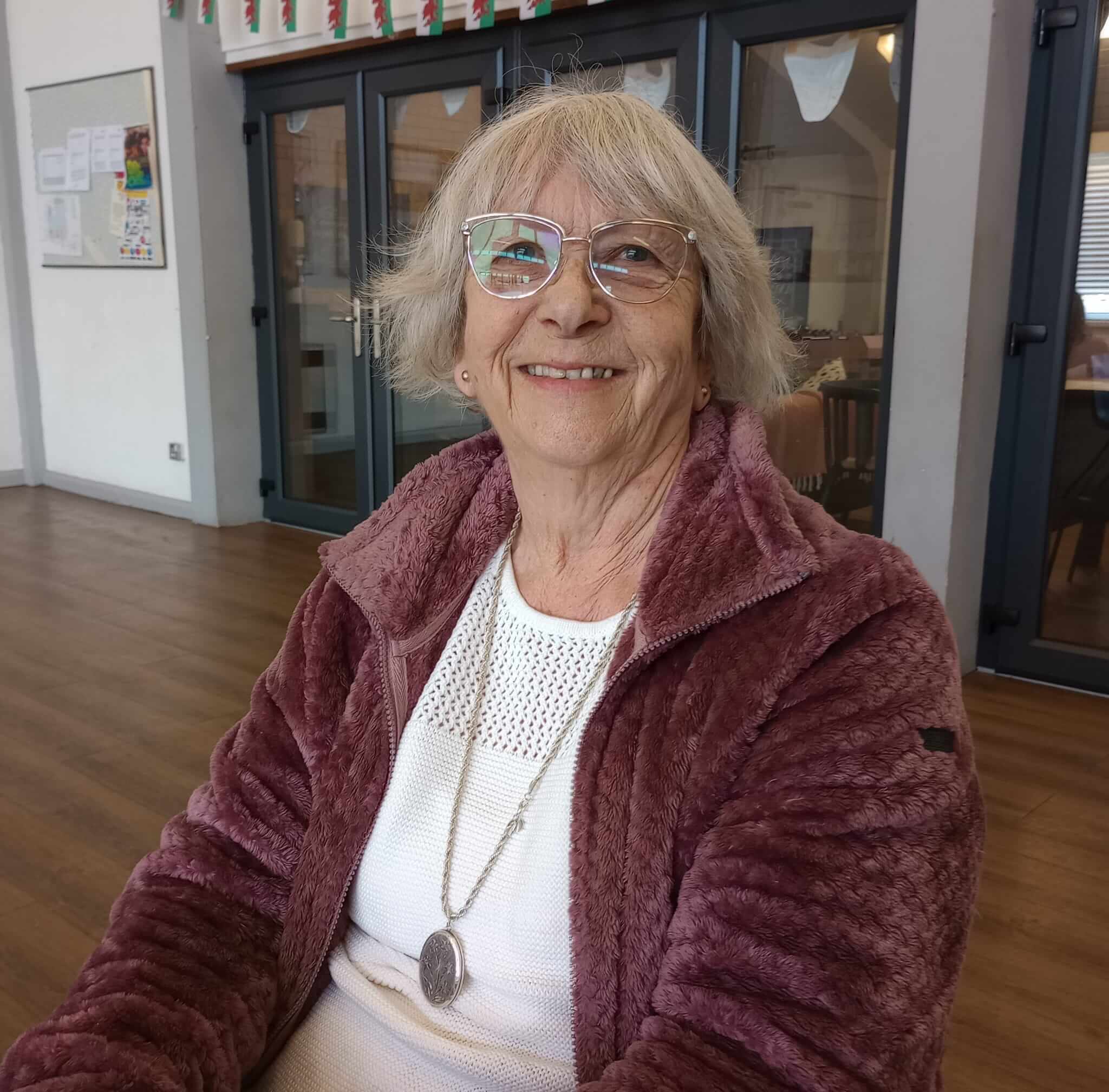 An elderly woman with glasses and a mauve jacket smiles while sitting indoors. Background includes glass doors. - Home Instead