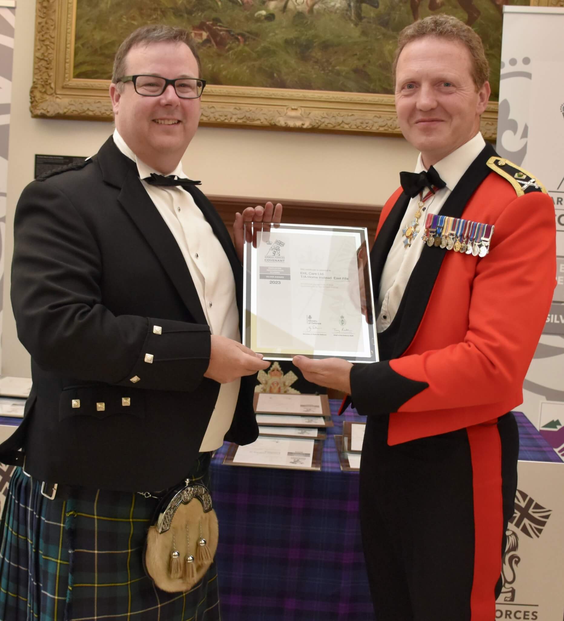 Two men in formal attire, one in a kilt and one in a military uniform, holding a framed certificate together. - Home Instead