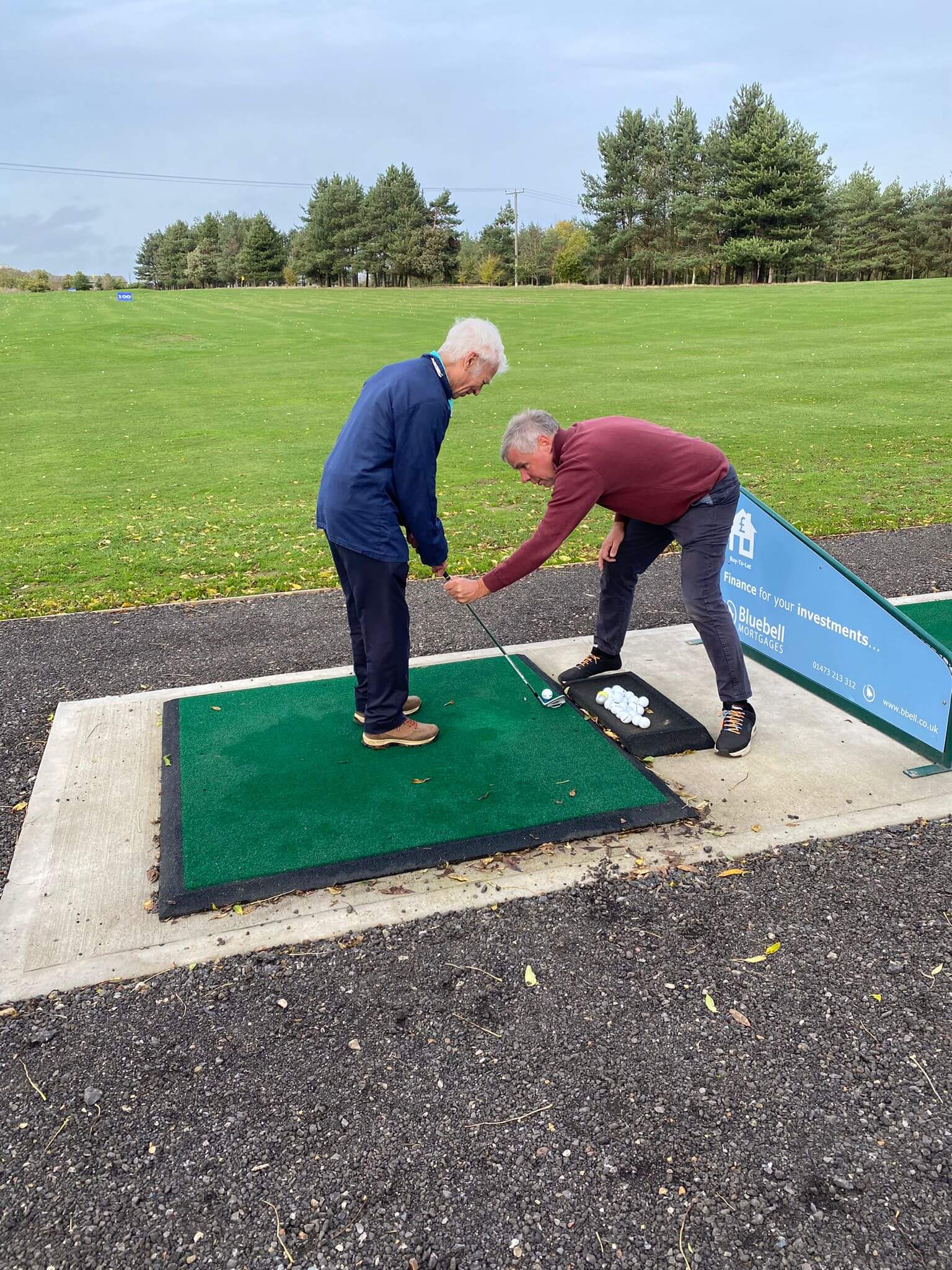 Two older men at a golf range, one helping the other with his golf swing on a practice mat. Trees and grass in the background. - Home Instead
