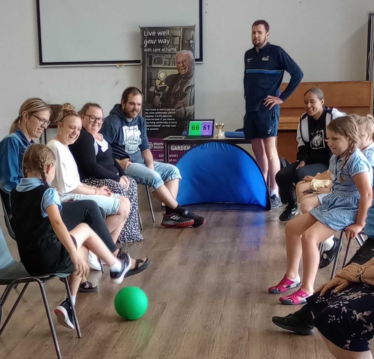 A group of adults and children sit in a circle passing a green ball. A person in sportswear stands behind them. - Home Instead