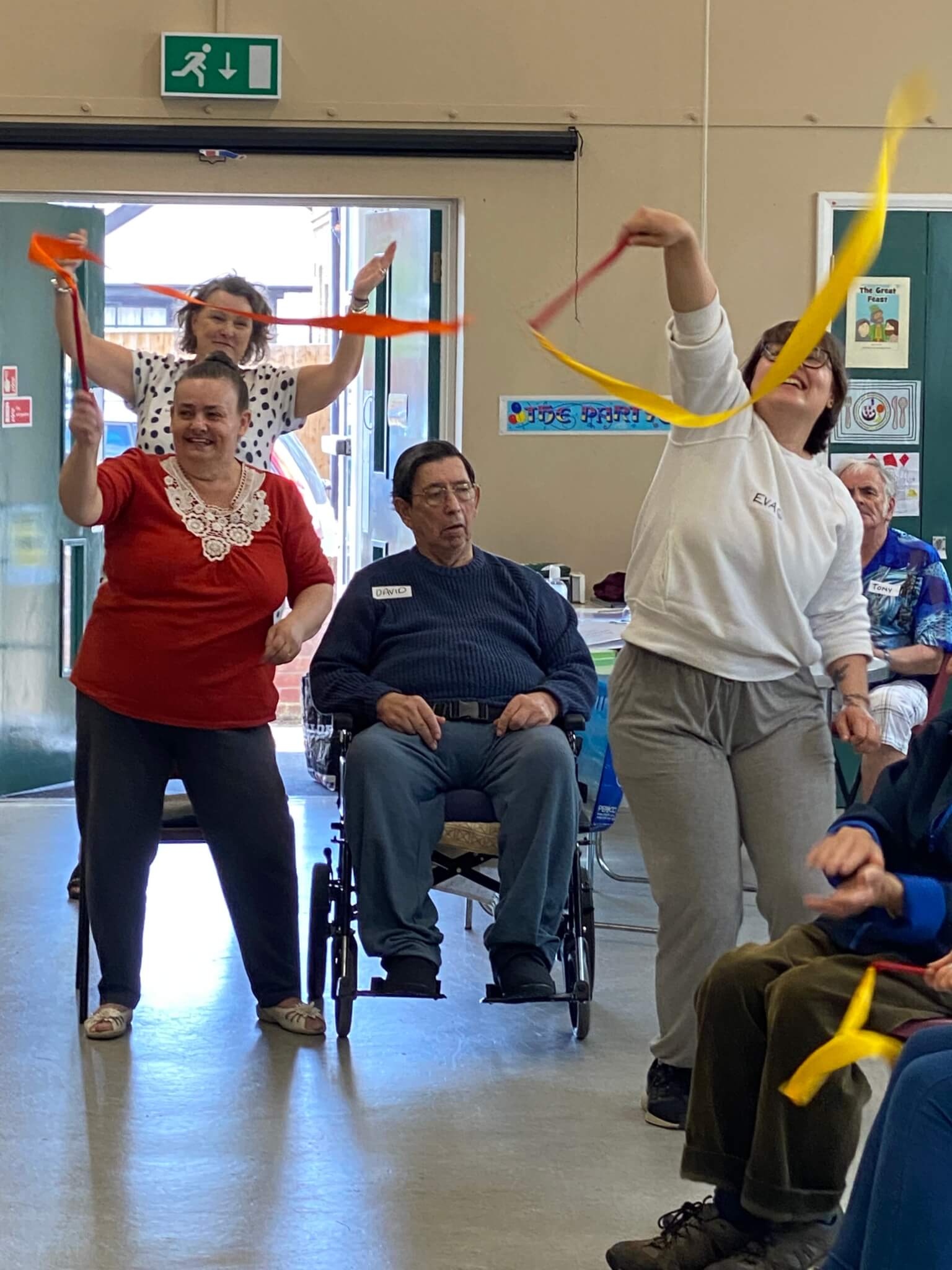 People are dancing with colorful streamers in a community hall, with seated elderly individuals watching and smiling. - Home Instead