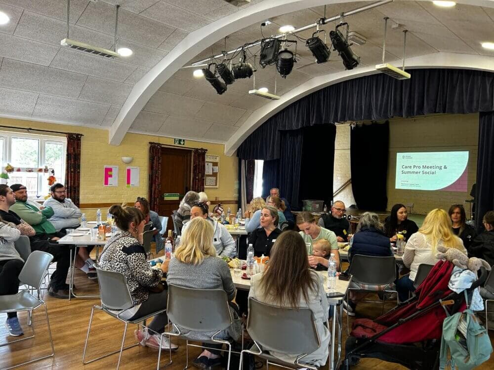 A group of people sitting at tables in a hall, attending a meeting and social event with a presentation screen. - Home Instead