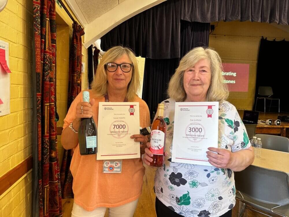 Two women hold certificates and bottles in a celebratory event in a decorated indoor space with a stage. - Home Instead