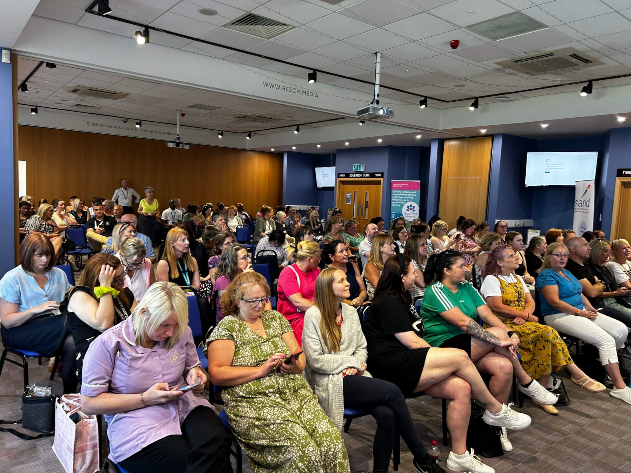 A large group of people seated in a conference room, attentively facing the front. - Home Instead
