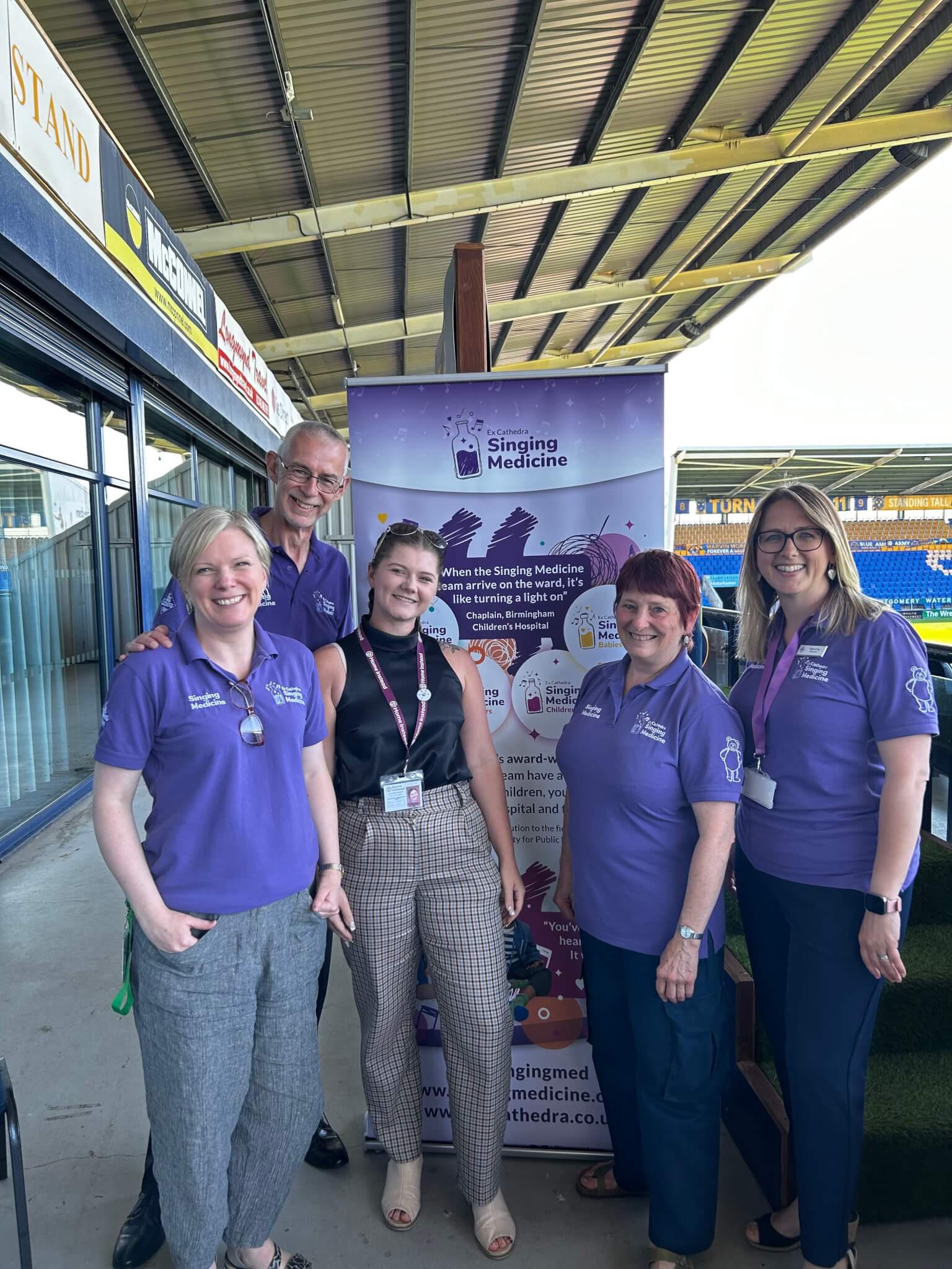 Five people standing and smiling in front of a "Singing Medicine" booth at a stadium. - Home Instead